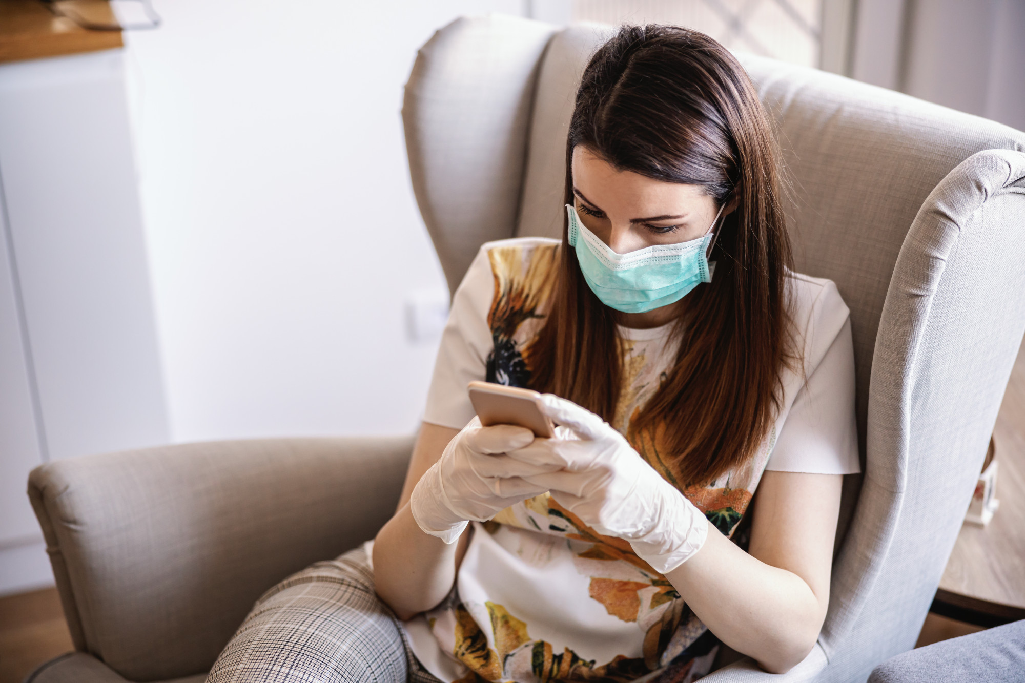 Jovem mulher em casa com máscara e luvas, olhando e digitando no smartphone. Ficar em casa. Pandemia Mundial.