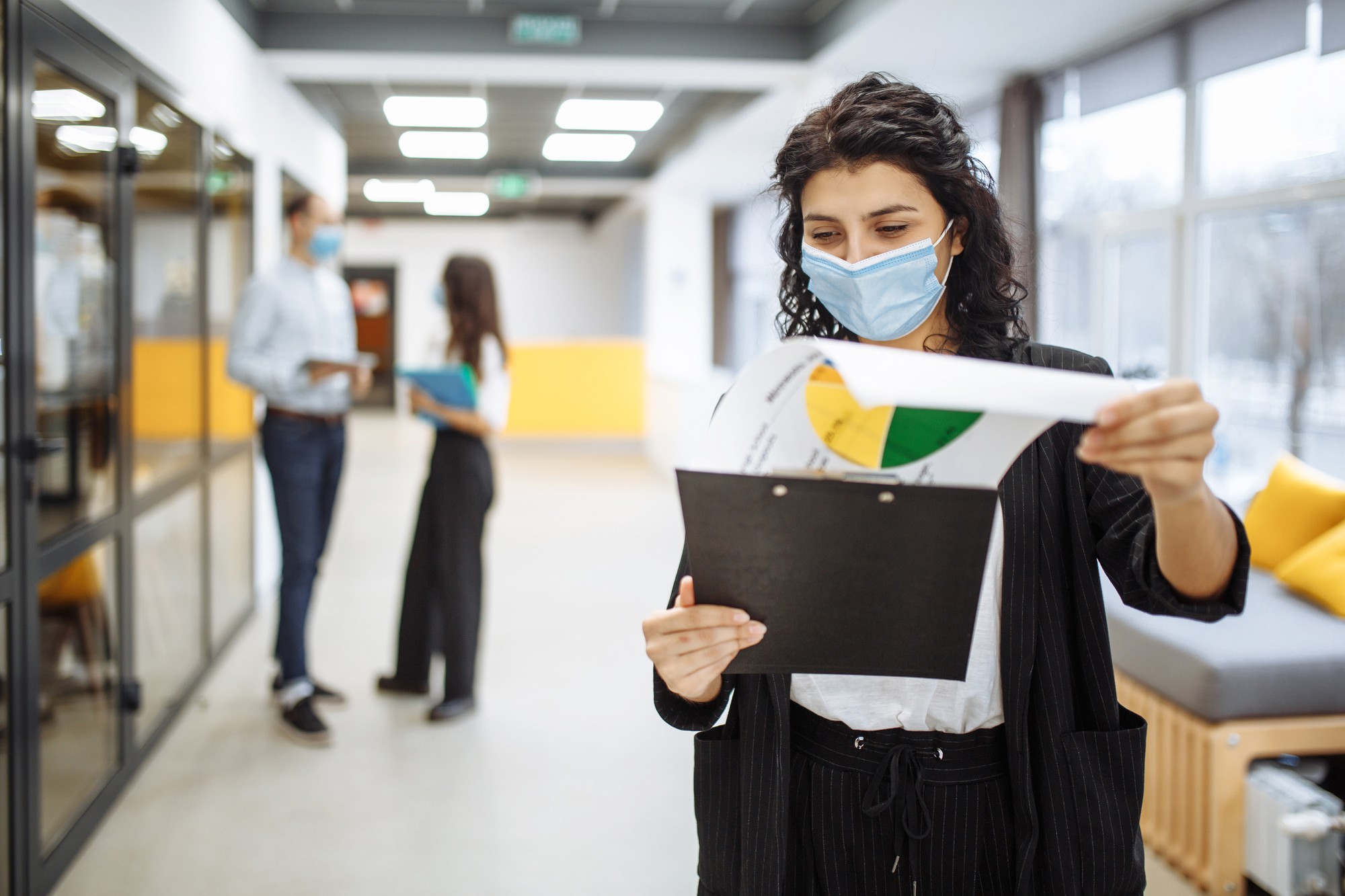 Retrato de uma jovem mulher de negócios usando uma máscara médica, verificando papéis, tendências e perspectivas de negócios em um corredor de escritório.