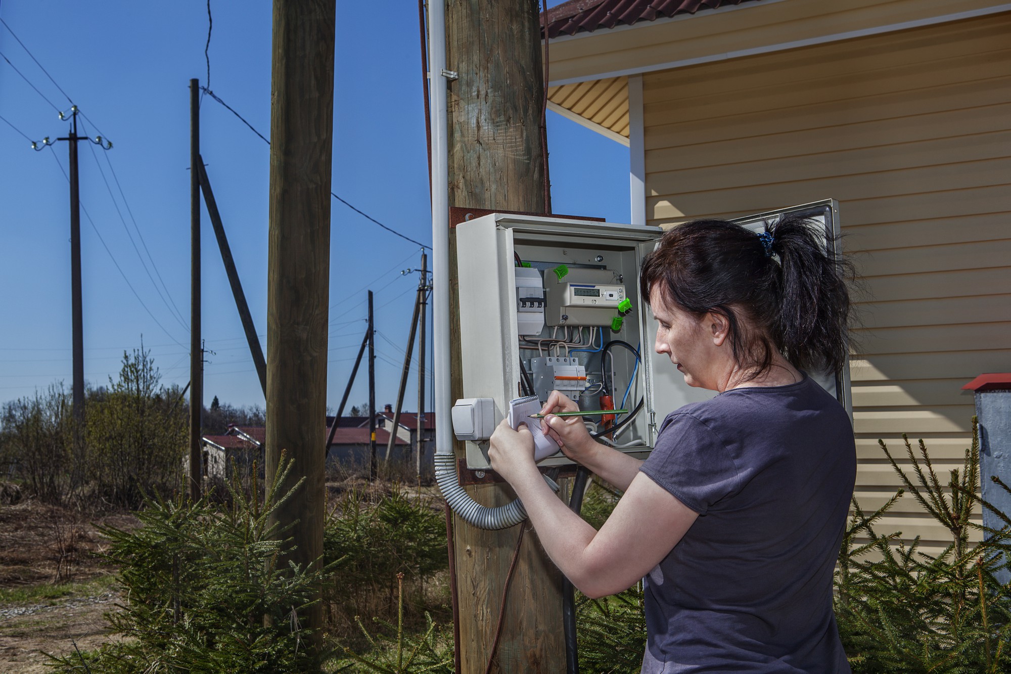 Mulher procurando dados do medidor elétrico em um painel montado em um poste de madeira perto de casa de fazenda na zona rural.