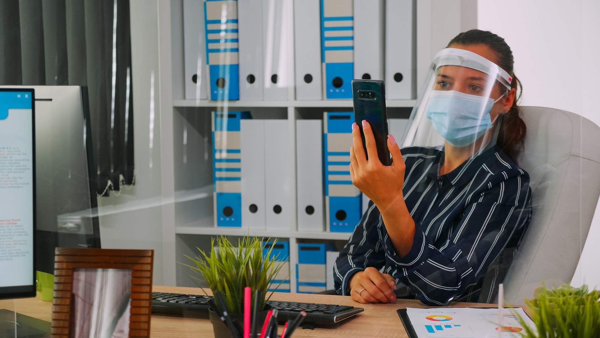 Empreendedor usando proteção facial contra coronavírus usando telefone para videoconferência falando com um colega de trabalho remotamente sentado em um novo prédio comercial normal. Mulher respeitando a distância social