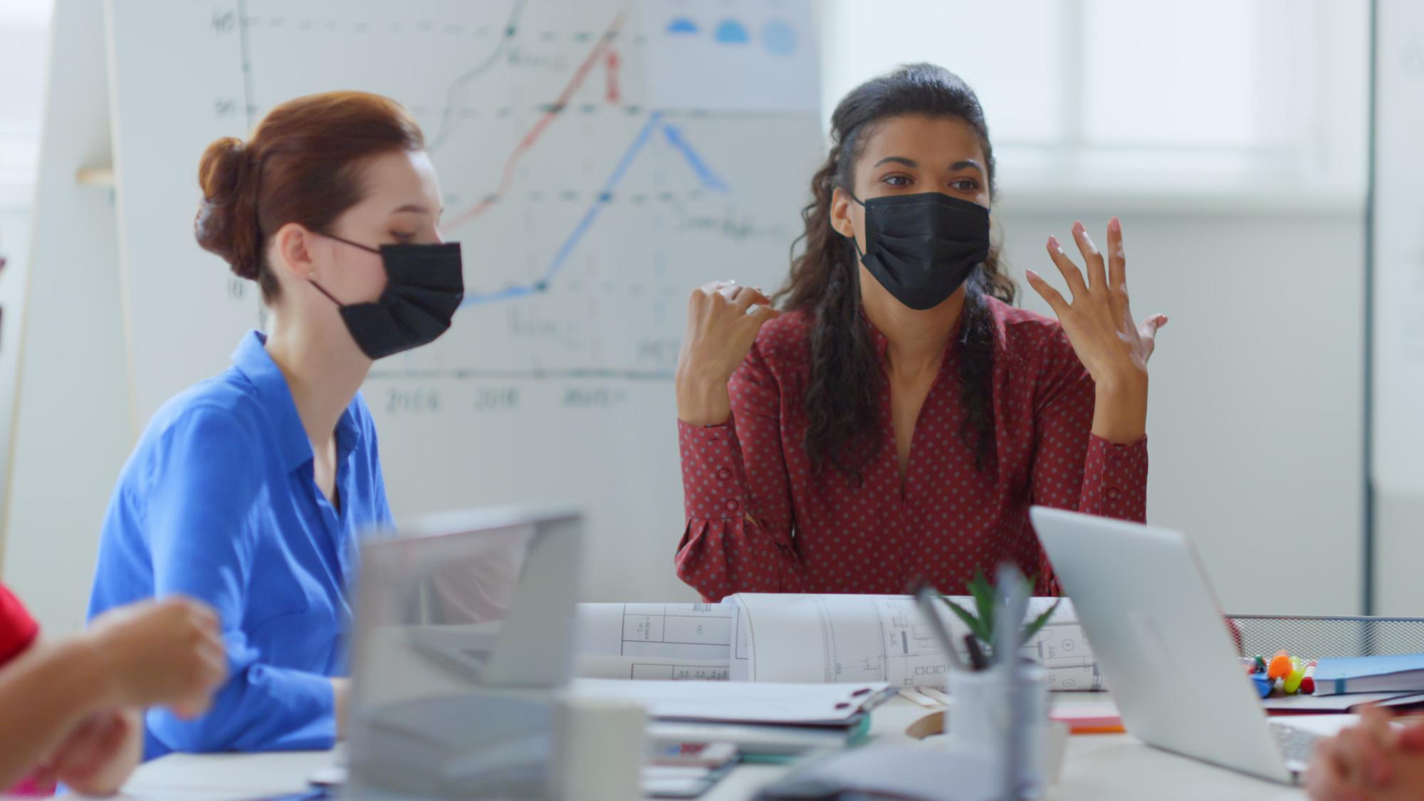 Mulheres usando máscaras protetoras estão sentadas em uma mesa com um gráfico atrás delas.