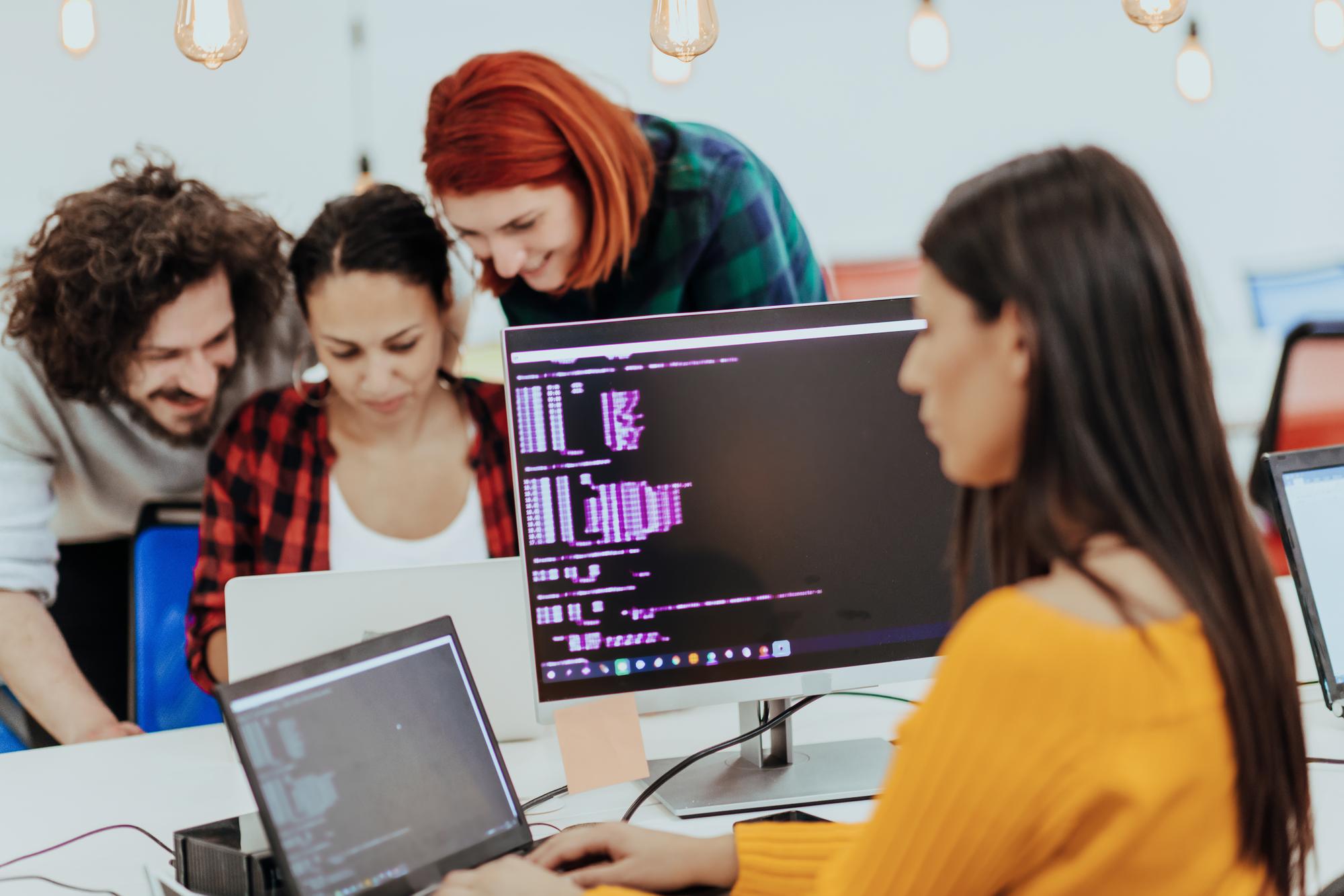 Equipe de desenvolvedores e profissionais de marketing colaborando em frente a uma tela com códigos e gráficos de performance