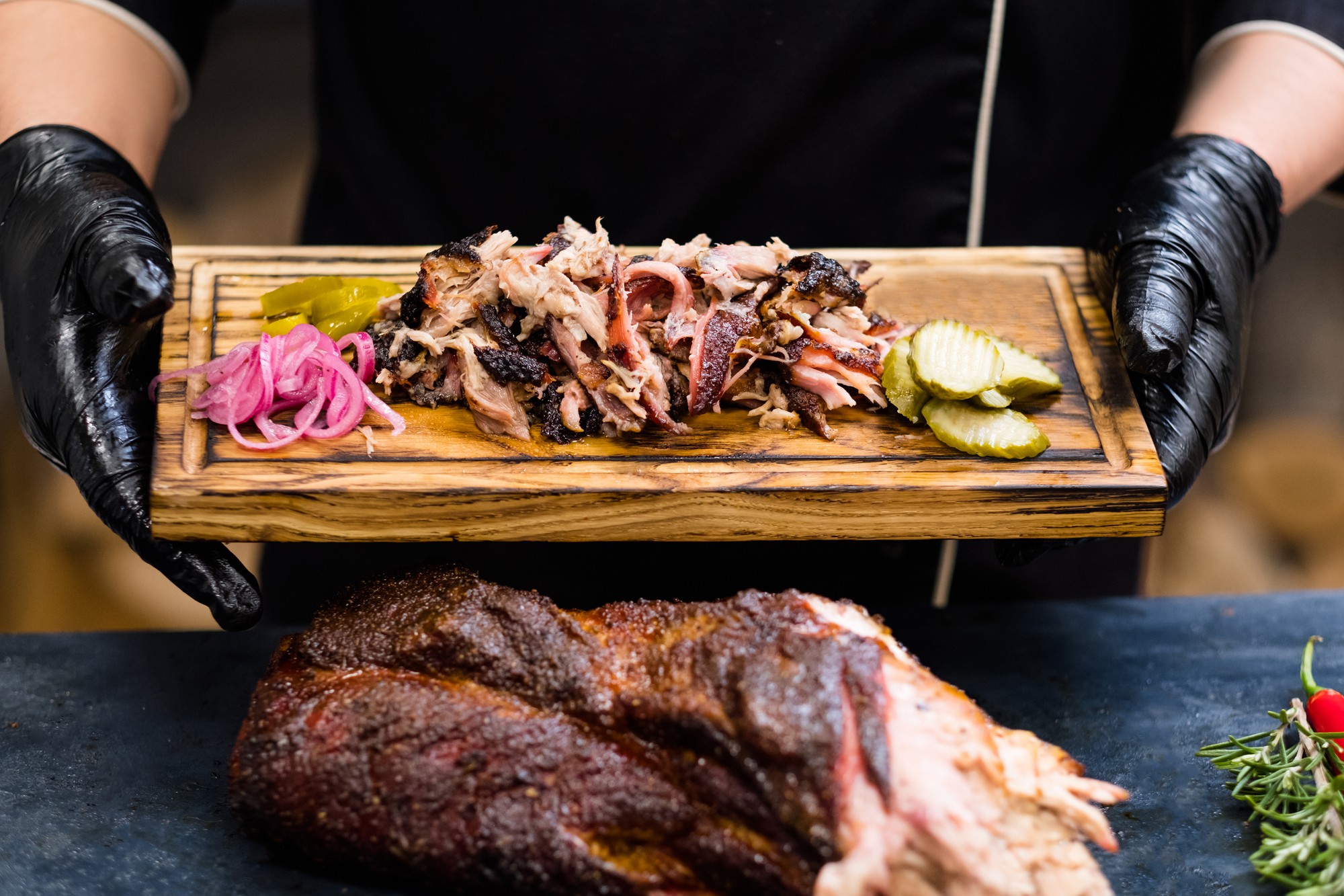 Cozinha de restaurante de grelhados. Chef mãos em luvas pretas de cozinha segurando carne de porco desfiada defumada com picles na placa de madeira.