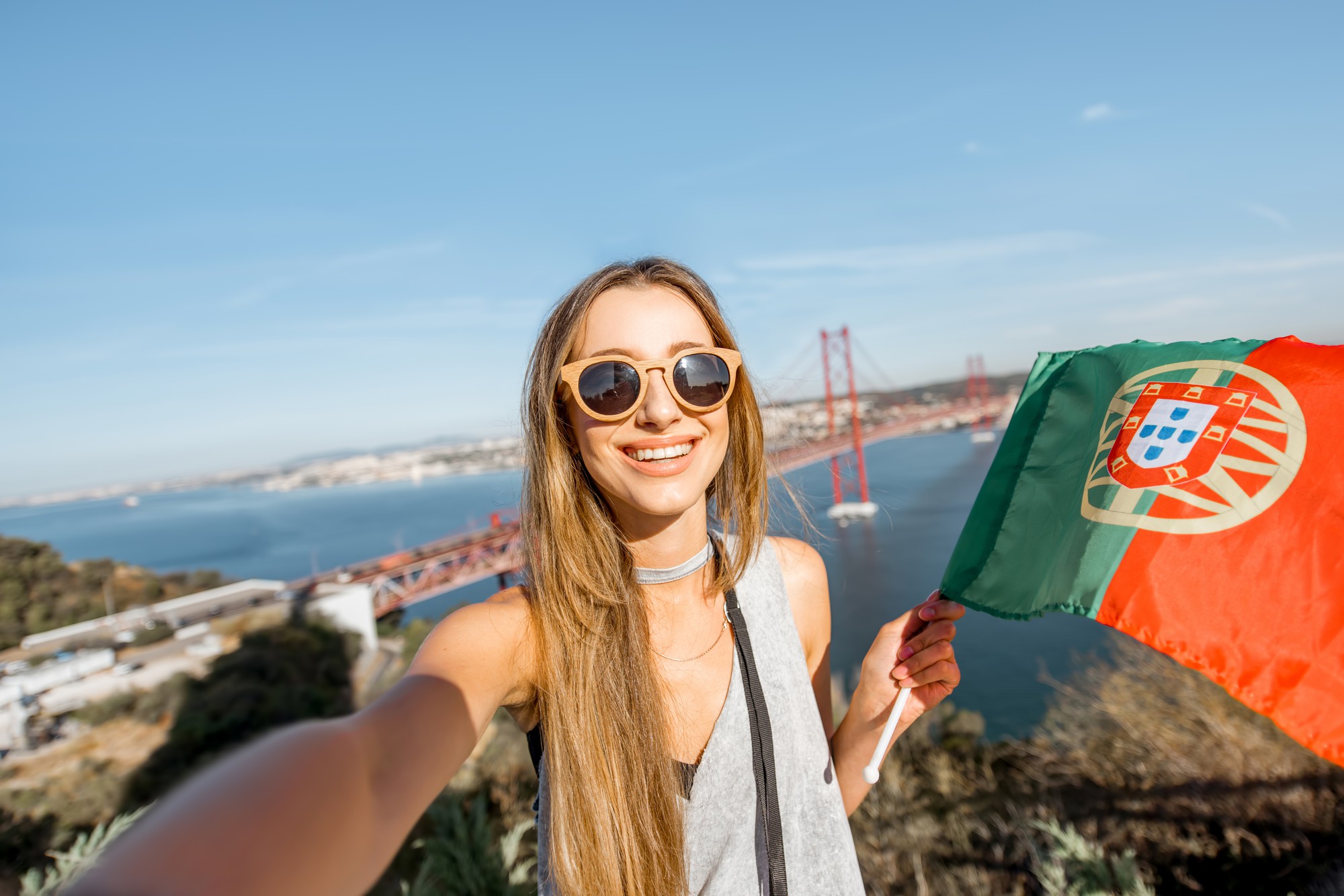 Jovem mulher fazendo selfie com a bandeira portuguesa no fundo da bela paisagem com a ponte e o rio em Lisboa, Portugal