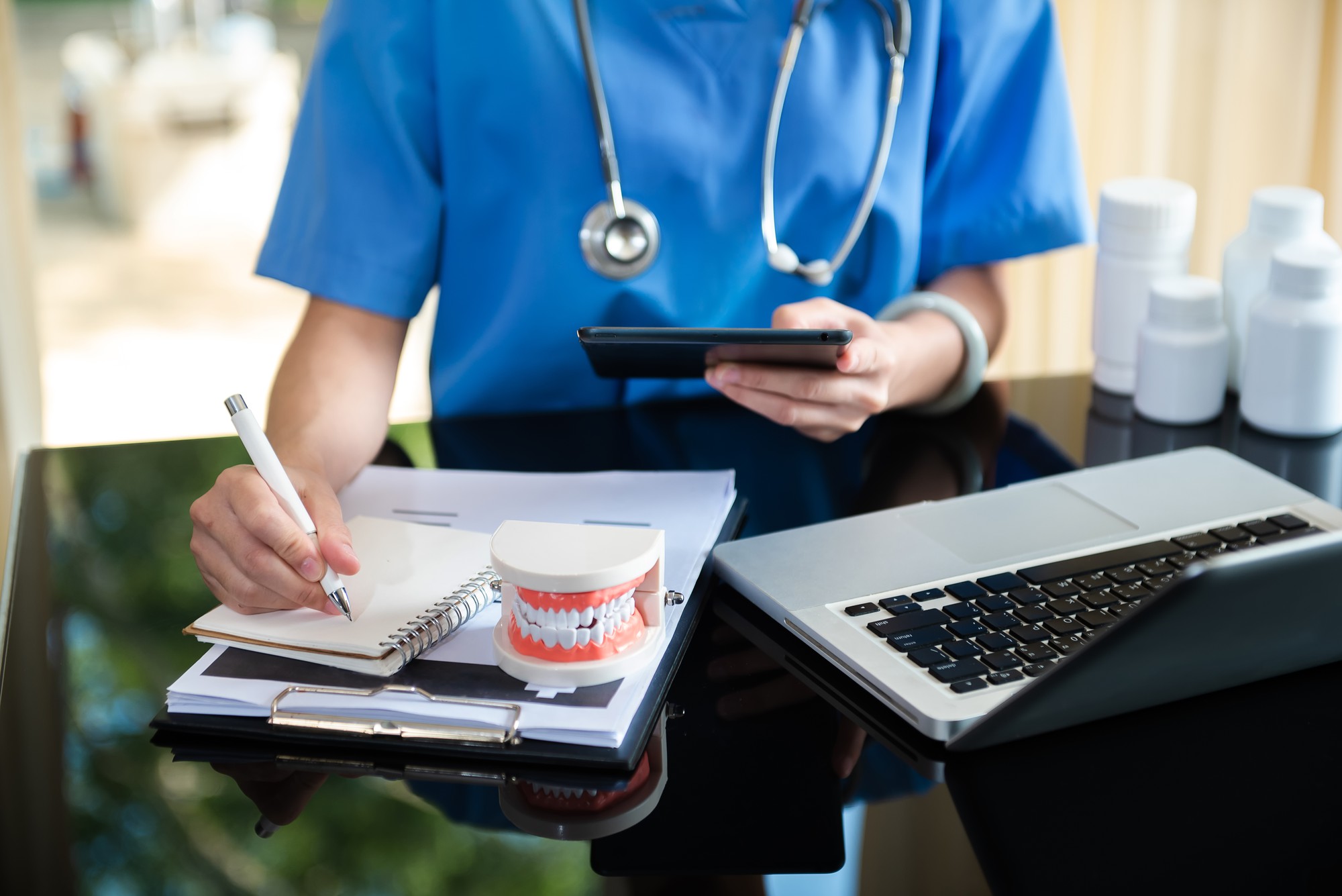 Dentista concentrado, sentado à mesa com modelo de dente de amostras de mandíbula e trabalhando com tablet e laptop em consultório odontológico clínica dentária profissional.