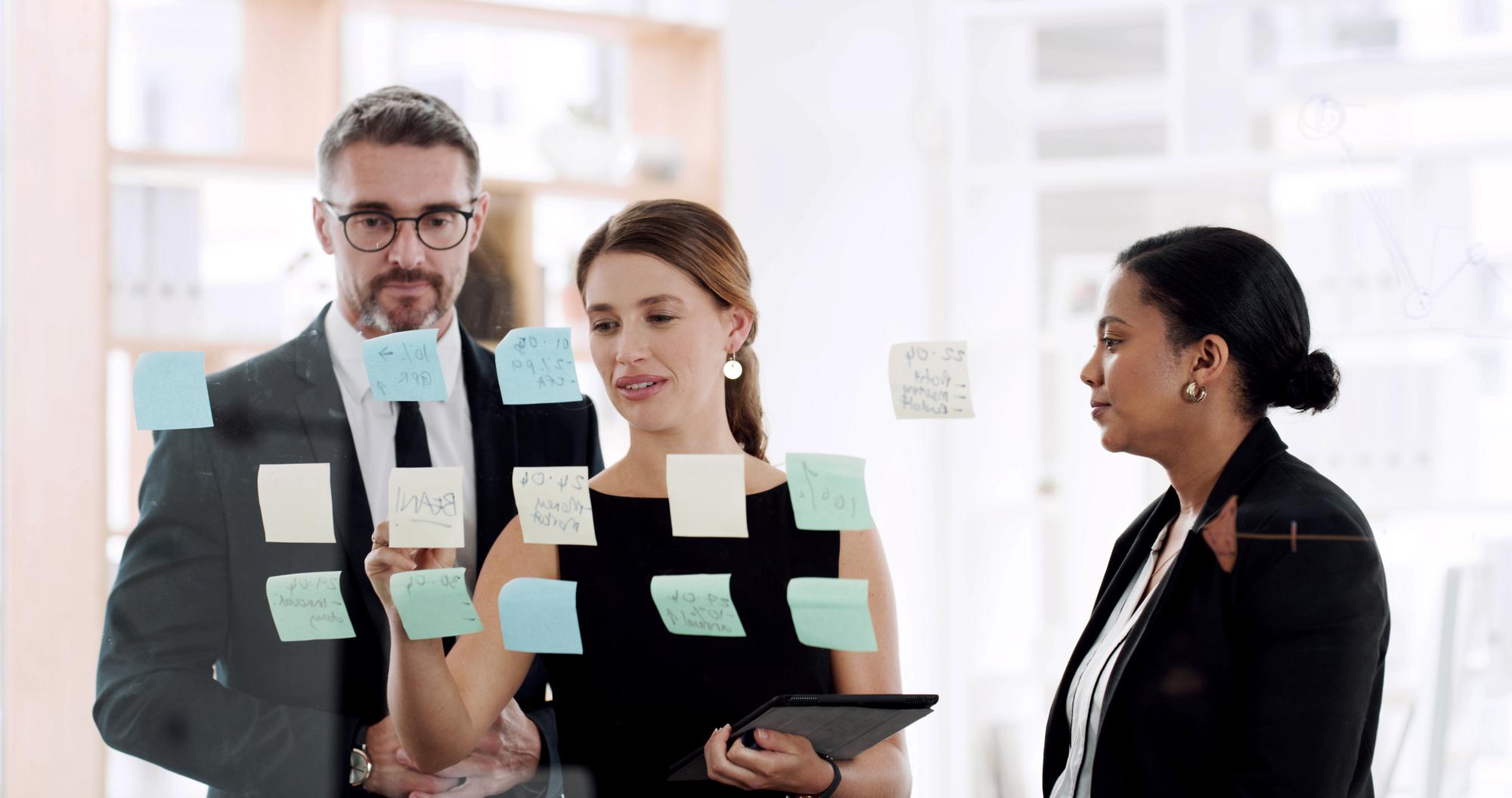 Pessoas de negócios planejando e escrevendo com post-it para reuniões ou tarefas agendadas no escritório. Grupo de funcionários ou quadro de vidro com tablet para ideias de estratégia de equipe ou brainstorming no local de trabalho